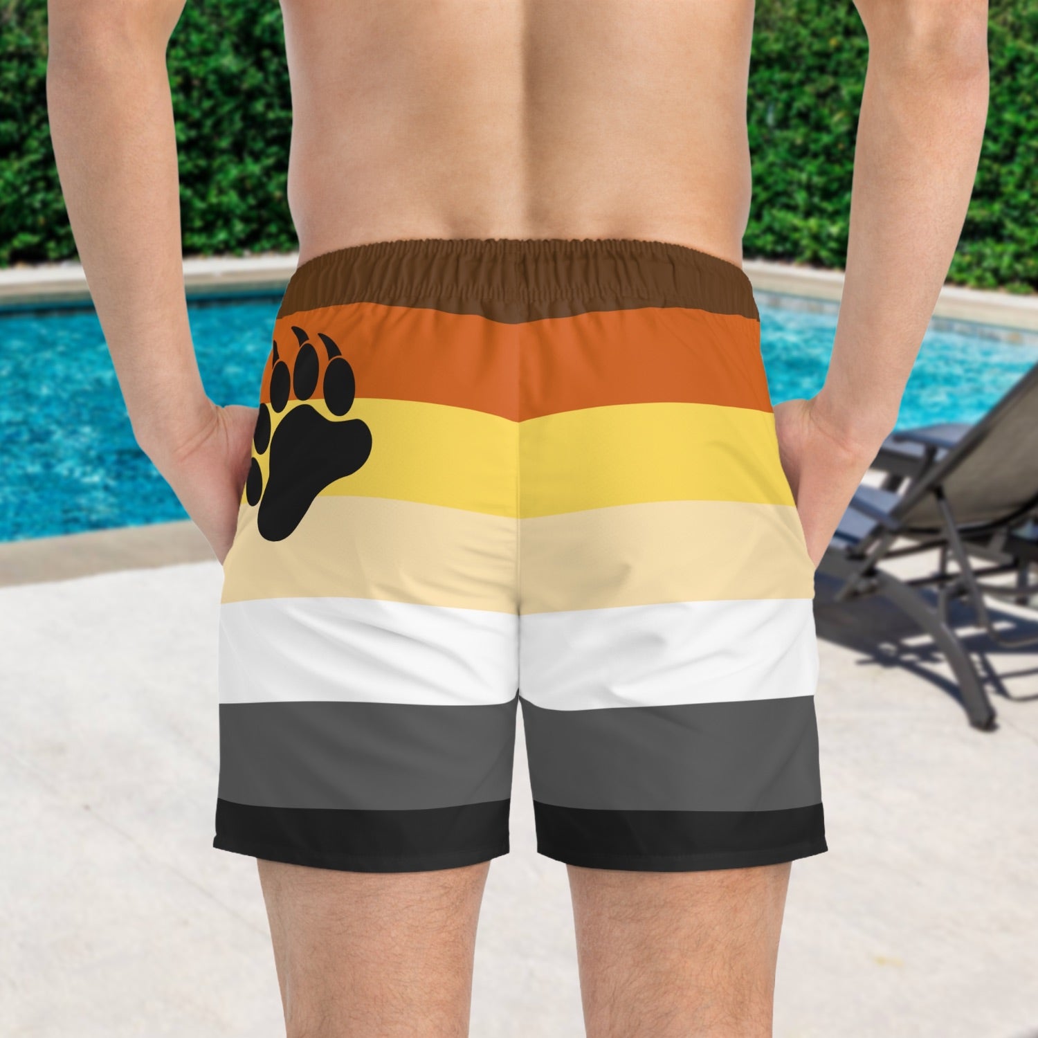 A person wearing striped pride flag swim shorts with a bear paw print design by a pool.