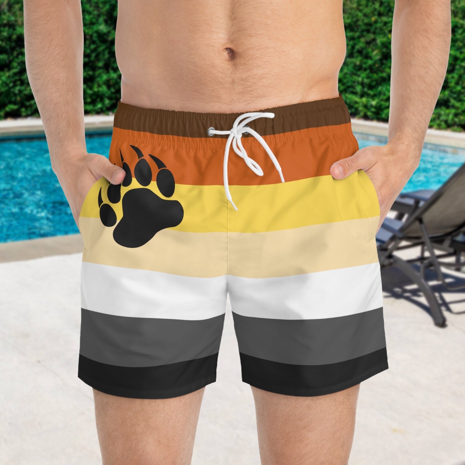 A person wearing striped pride flag swim shorts with a bear paw print design by a pool.