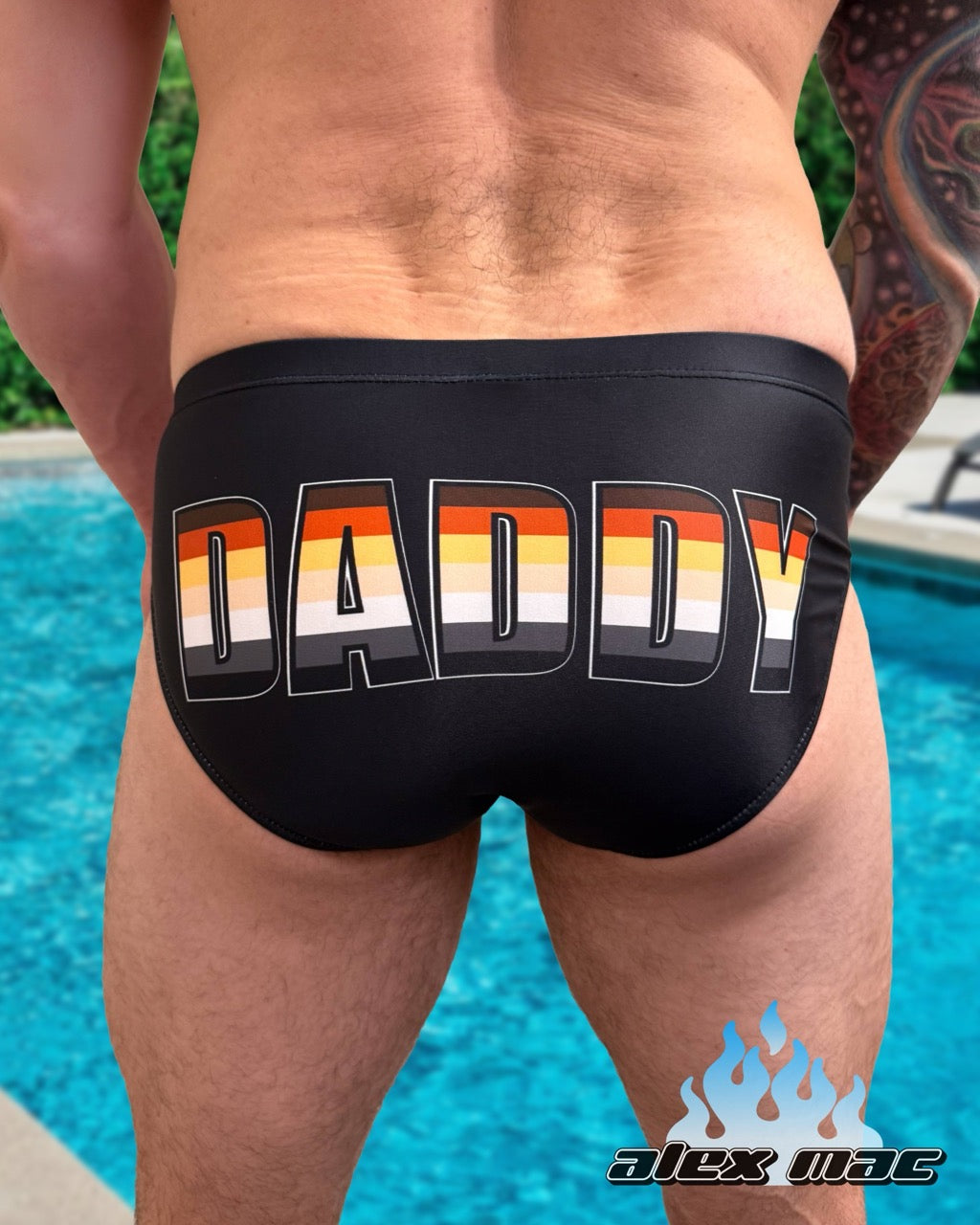 Black speedo swim brief with 'DADDY' text in bear pride flag rainbow colors. The Swim trunks are worn by a muscular, tattooed man by a pool.